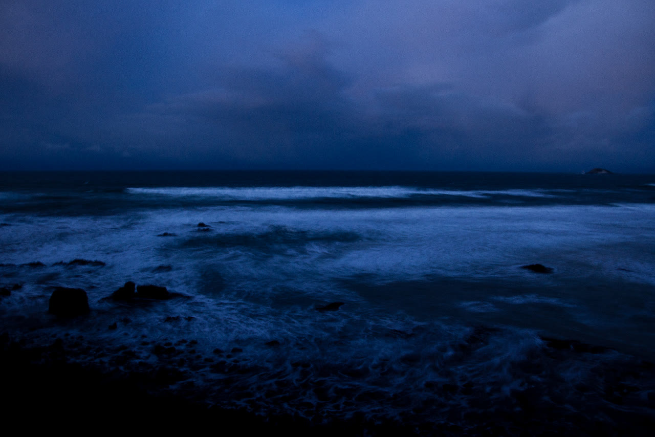 Dusk over a rocky shoreline. Waves roll in white across dark water toward black rocks in the foreground, the whole scene washed in deep blue and violet. Low cloud fills the sky, and a small island sits dark on the horizon at the right.
