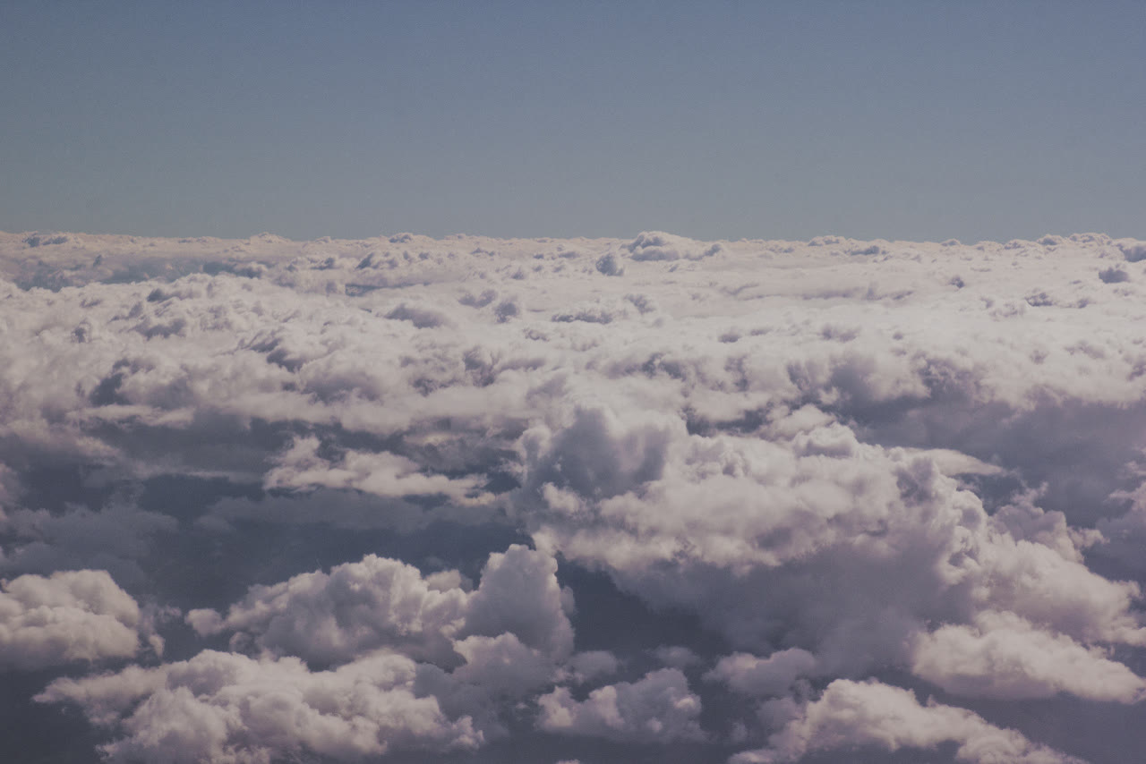 Looking down from high above, a field of white and lavender-grey cloud stretches to a flat horizon. The cloud tops pile in soft, rounded heaps with shadowed hollows between them, beneath a clear, pale blue sky.