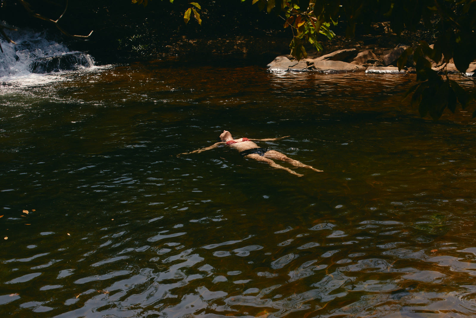A person floats on their back in a dark, tea-brown pool, arms spread wide and face turned up to the sun. A small waterfall spills white over rocks at the upper left, and overhanging leaves frame the top of the picture.