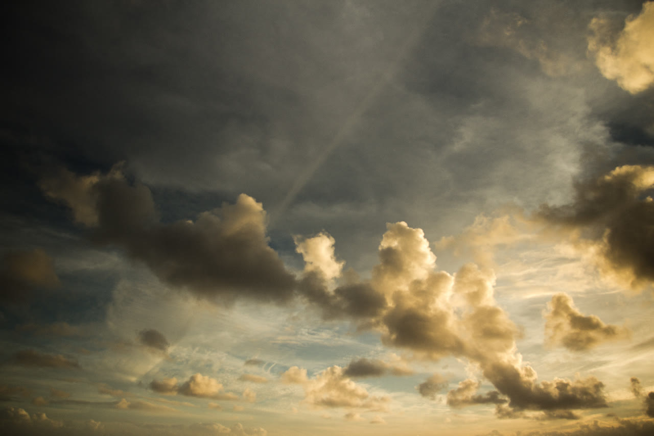 A wide sky of heavy cloud at sunset. Dark slate-grey masses drift across the top of the frame, while the lower clouds are lit gold and cream along their edges. Thin, pale streaks of high cloud cross a faded blue.