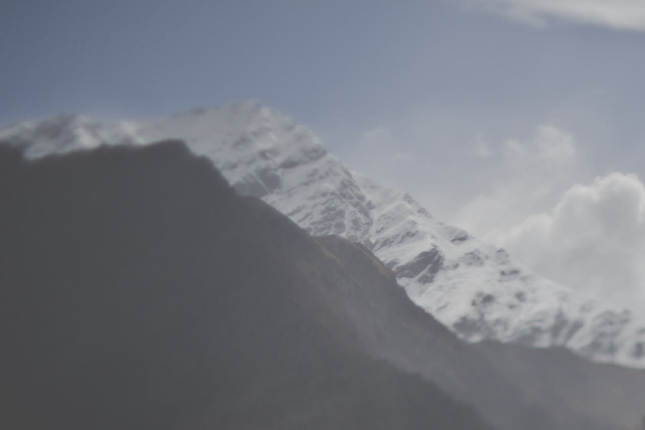 A snow-covered mountain peak, softly out of focus, rises behind a darker ridge in the foreground. The snow is bright and broken by grey rock, under a pale, cool blue sky with white cloud gathering at the right.